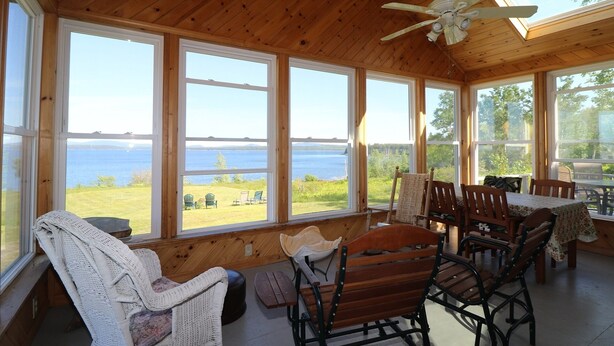 A wooden cabin with large windows overlooking a lake, furnished with wicker chairs and a wooden table.
