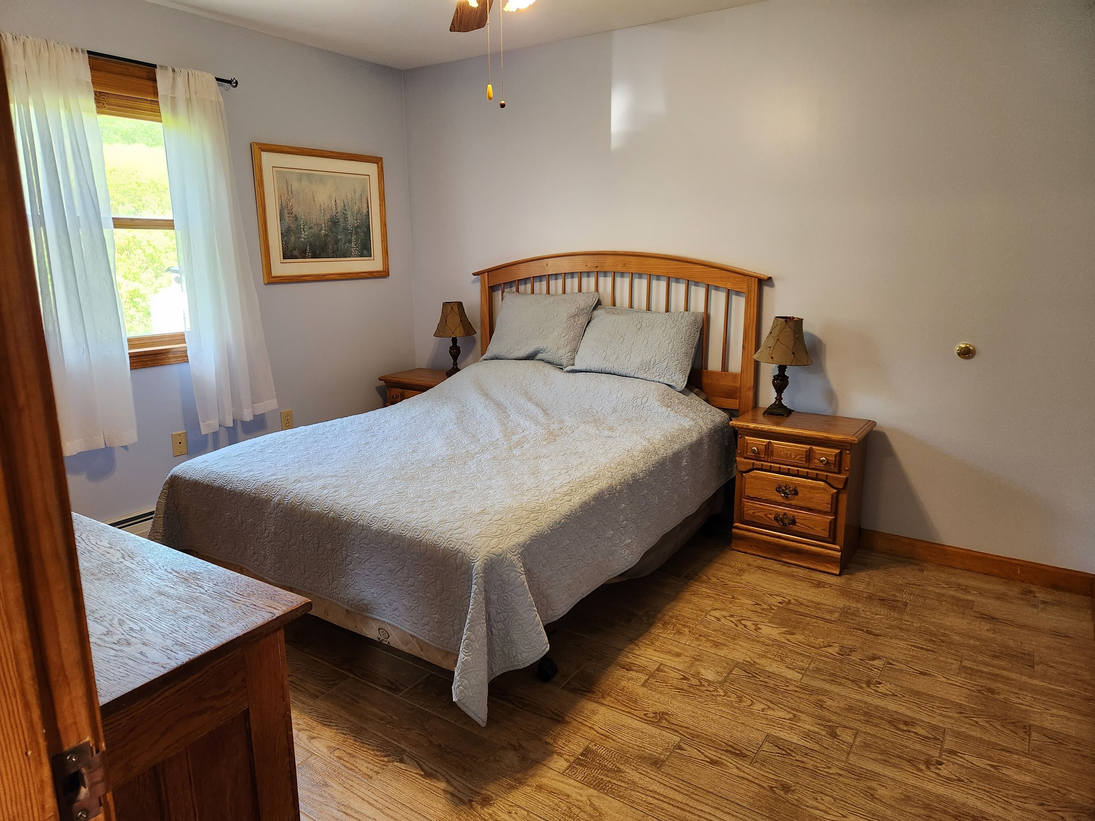 Une chambre avec un lit en bois, deux tables de chevet avec des lampes, une commode, un tableau encadré au mur et une fenêtre avec des rideaux.