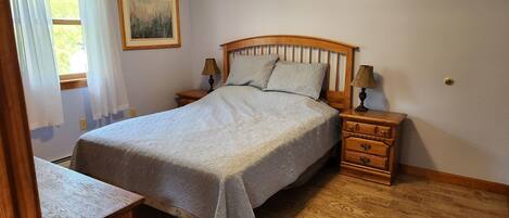 A bedroom with a wooden bed, two bedside tables with lamps, a nightstand, a framed picture on the wall, and a window with curtains.