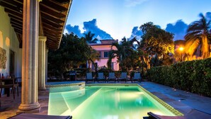Ein Swimmingpool mit Liegestühlen, einer überdachten Sitzgelegenheit und einem Gebäude im Hintergrund.