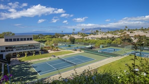 A tennis court complex with multiple courts, a building with a glass facade, and a mountainous backdrop.