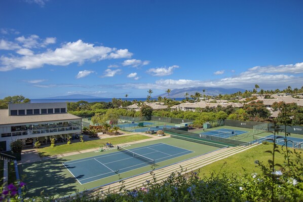 Un complexe de courts de tennis comprenant plusieurs terrains, un bâtiment à la façade en verre et un paysage montagneux en arrière-plan.