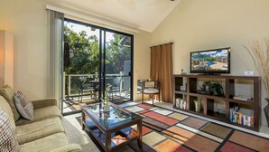 A living room with a glass door leading to a balcony, a television on a wooden stand, and a bookshelf filled with books.