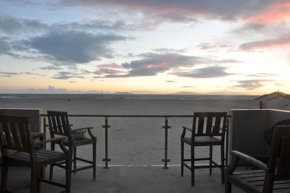 Un balcón con dos sillas de madera y una mesita pequeña, con vistas a una playa y un atardecer al fondo.