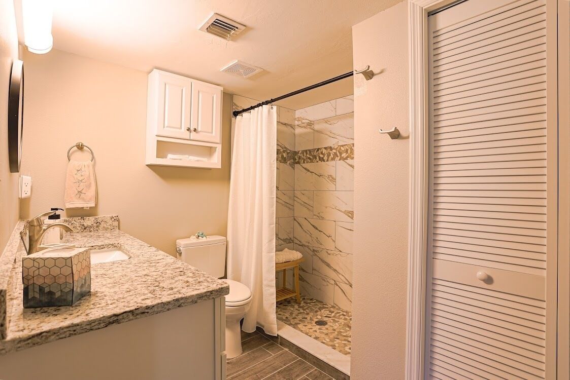 A bathroom with a marble shower, a toilet, a sink with a soap dispenser, and a cabinet above the sink.