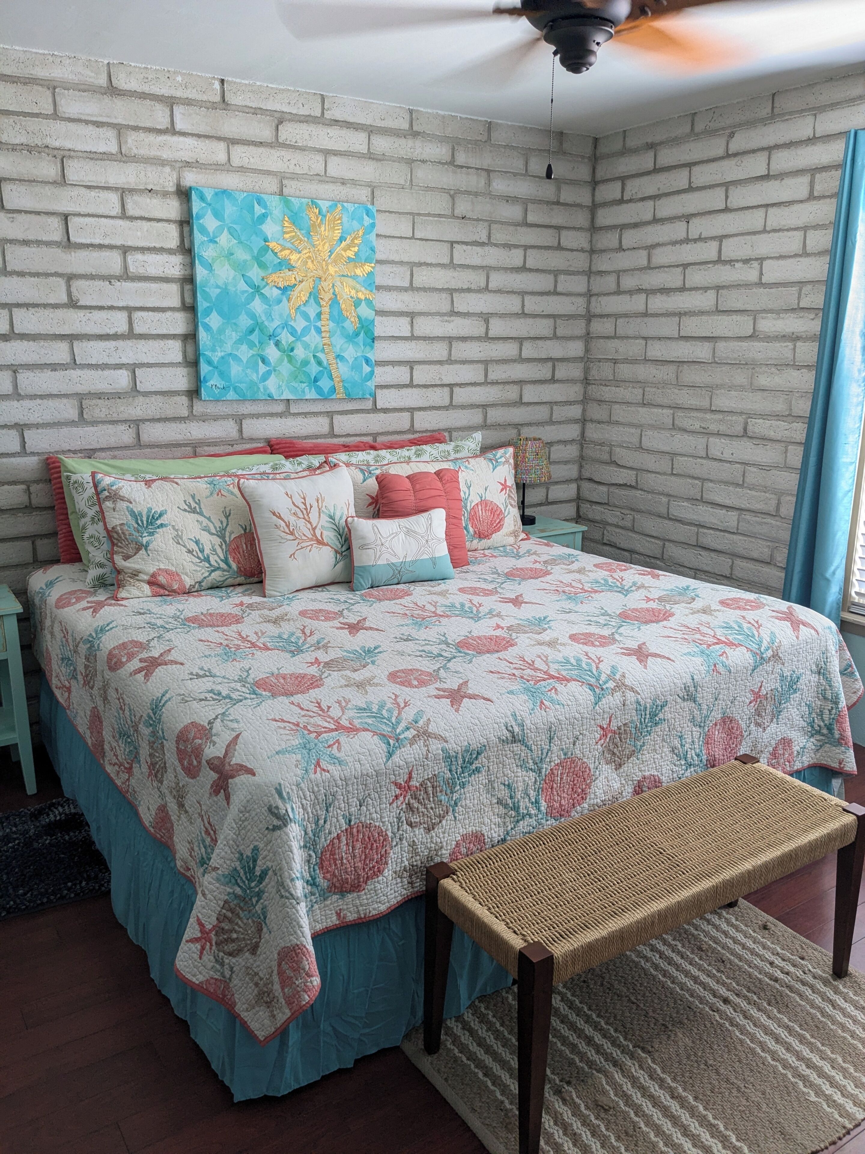 Ein Schlafzimmer mit Backsteinwand, einem Bett mit floralem Bettzeug, einer gewobenen Bank und einem Deckenventilator.