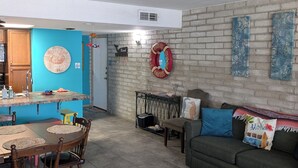 Un comedor con una mesa redonda y sillas, una cocina con pared azul y una puerta, una pared de ladrillo con obras de arte, y un ventilador de techo.