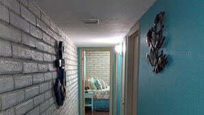 Un pasillo con pared de ladrillo, un felpudo y una cama frente a una pared azul.