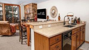 A kitchen with a stone fireplace, a large island with a sink, wooden cabinets, and a dining area with a leather couch and chairs. - Townsend Place 205 | 2BR Ski-In/Ski-Out Condo, Hot Tub (Avon)