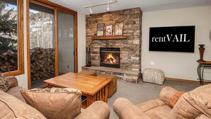 A cozy living room with a stone fireplace, comfortable seating, and a large TV mounted on the wall. - Townsend Place 205 | 2BR Ski-In/Ski-Out Condo, Hot Tub (Avon)