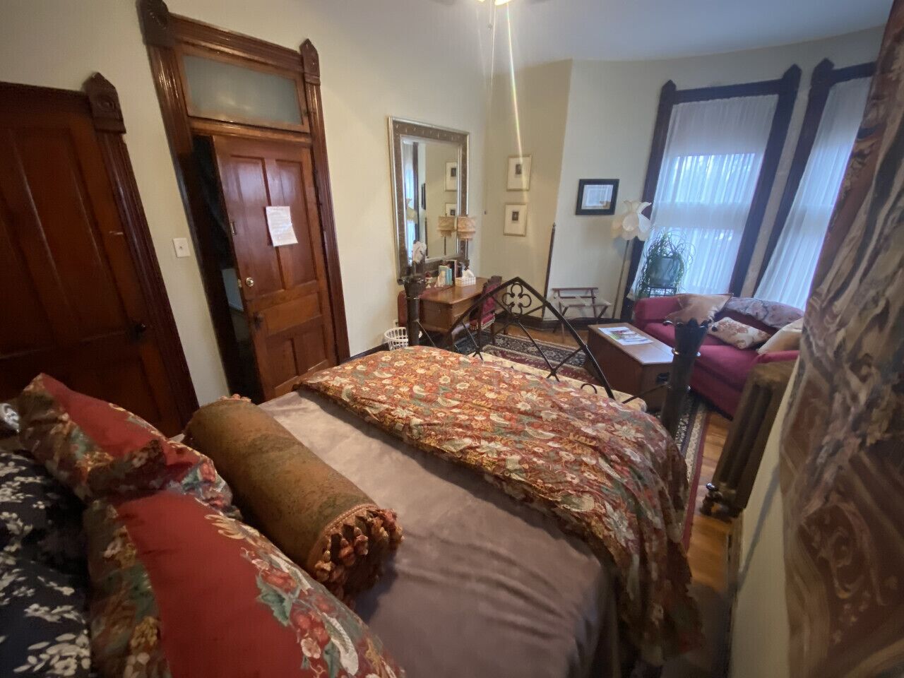 A bedroom with a large bed, a wooden door, a mirror, and a window with curtains.