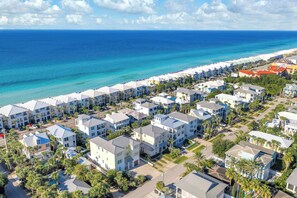 A coastal residential area with beachfront properties, including multi-story buildings with balconies, a beach with umbrellas, and clear blue skies.