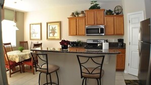 A kitchen with wooden cabinets, a microwave, and a refrigerator. There is a dining table with chairs and a bar area with stools.