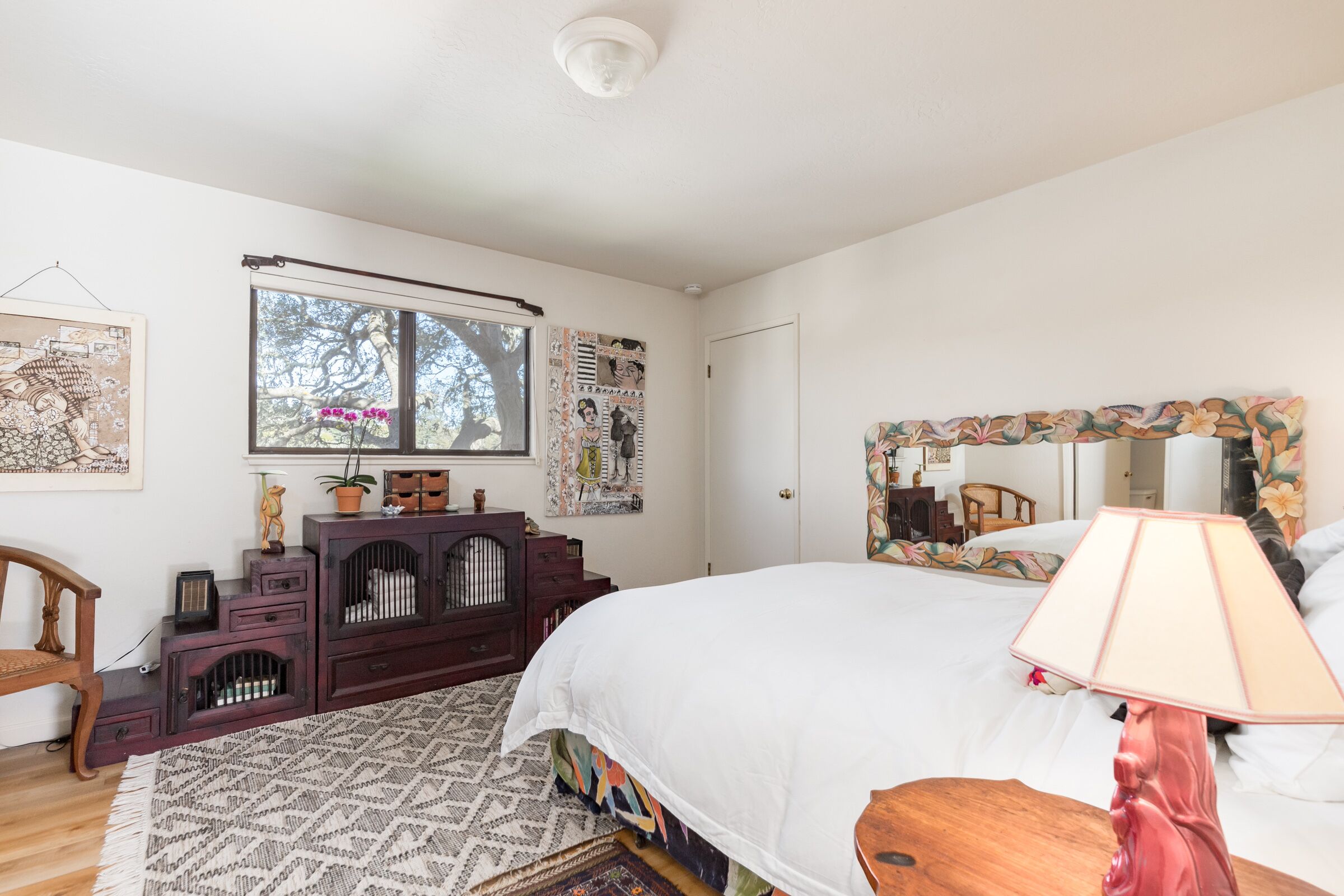 Ein Schlafzimmer mit einem großen Bett, einem Schminktisch, einem Spiegel und einem Fenster mit Blick auf Bäume.
