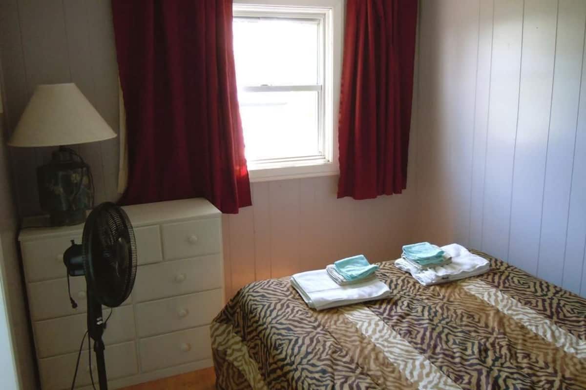Ein Schlafzimmer mit einem Bett, einem Ventilator, einer Kommode und einem Fenster mit Vorhängen.
