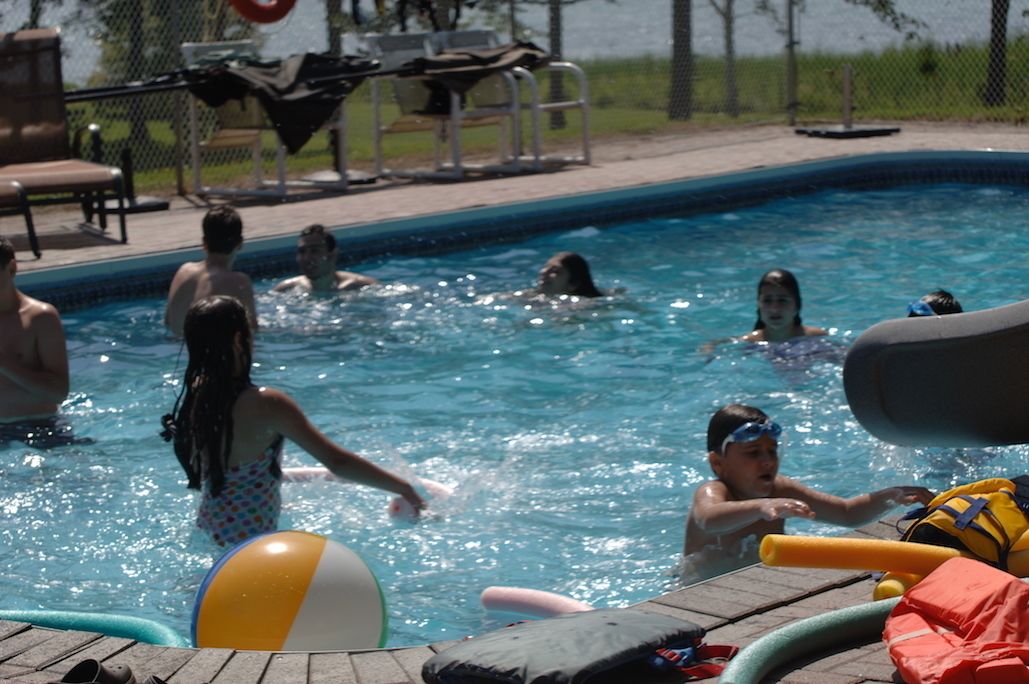 Una piscina con gente disfrutando, rodeada de sillas de descanso y una valla.