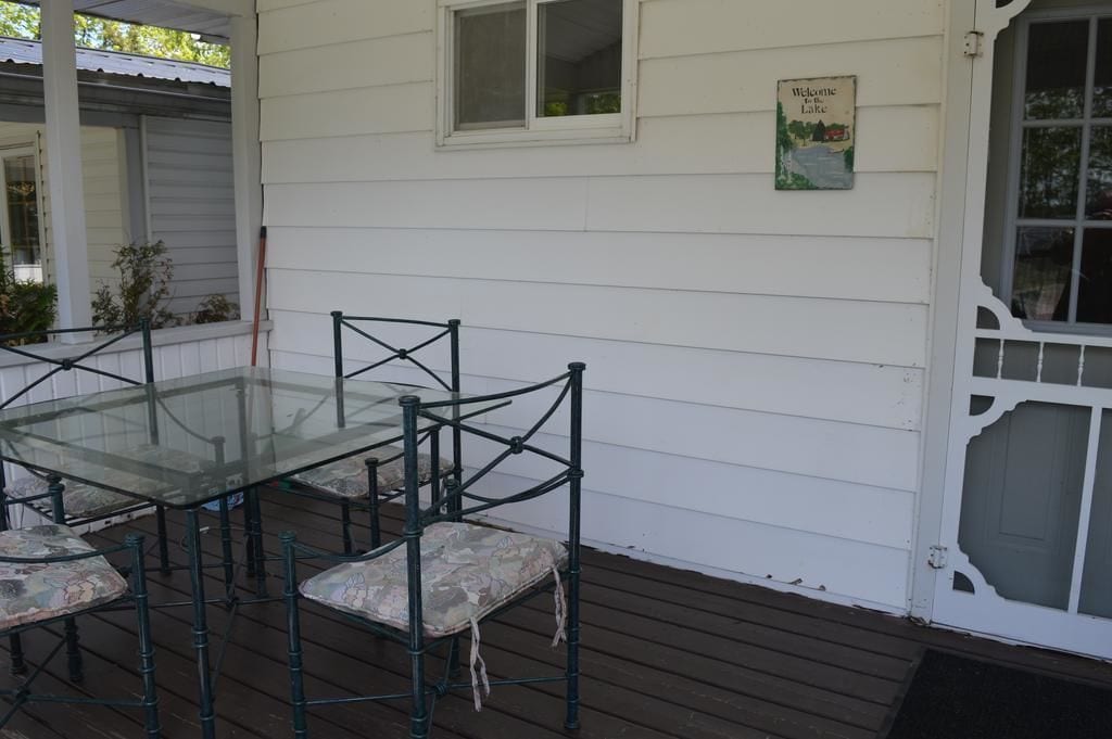 A glass-top table with chairs on a wooden deck, a white door with a glass panel, and a welcome sign on the wall.