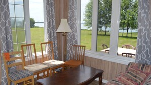Una habitación con una mesa de madera, cuatro sillas, un sofá y una lámpara. Amplias ventanas ofrecen vistas a un área verde y árboles.