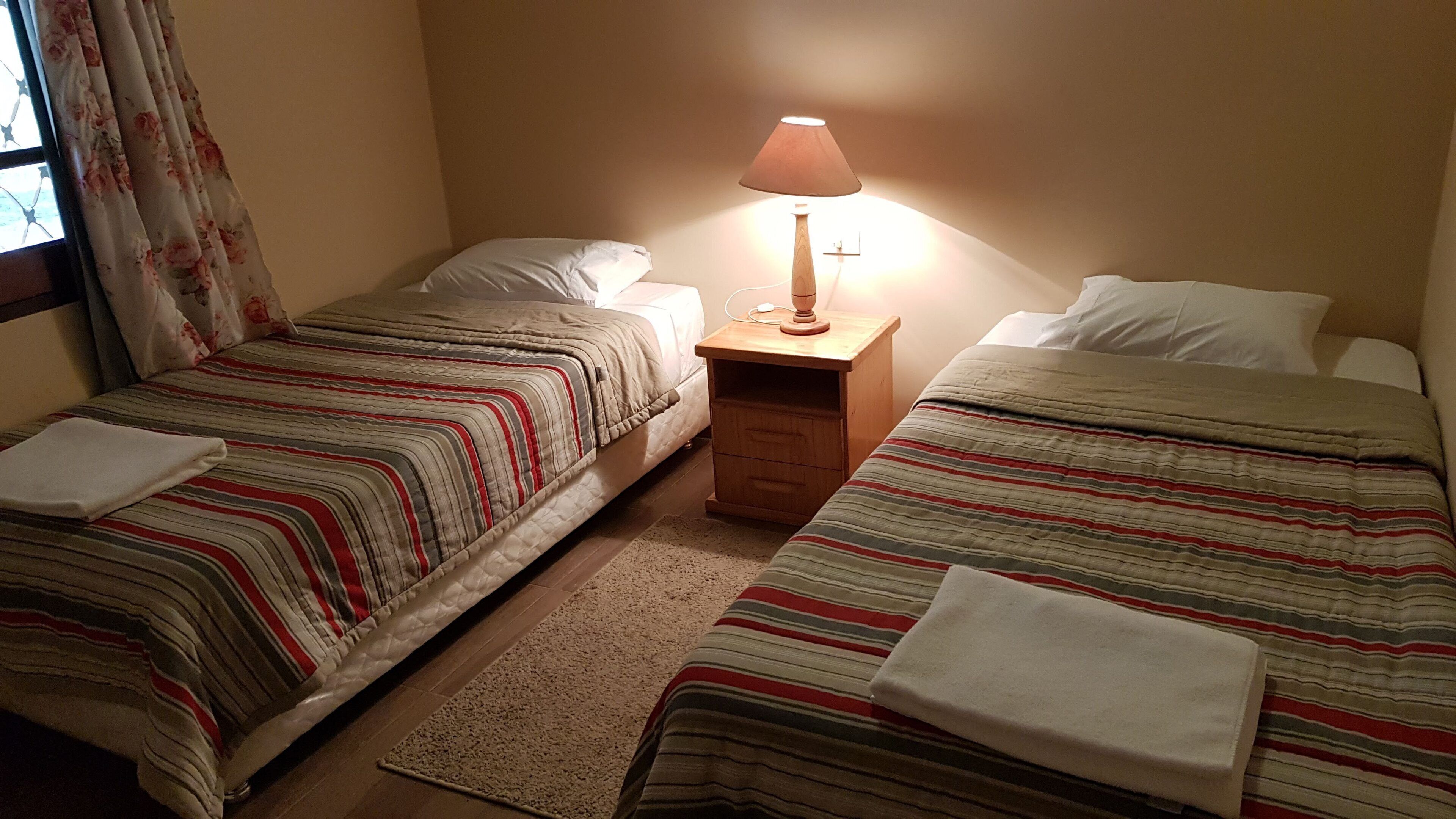 A room with two single beds, each with striped bedding and pillows, a wooden nightstand with a lamp, and a window with floral curtains.