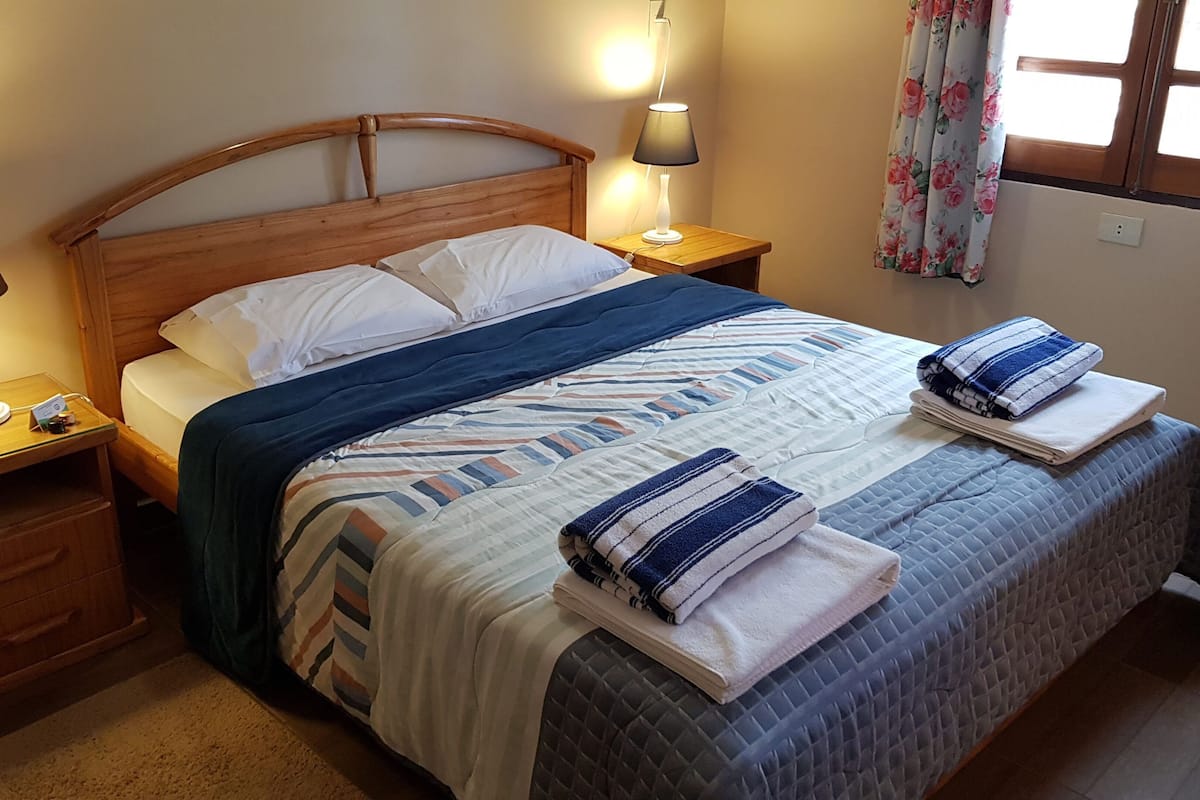 Una cama con cabecera de madera, dos mesitas de noche con lámparas, un ventanal con cortinas florales y una pequeña mesita de noche con una lámpara.