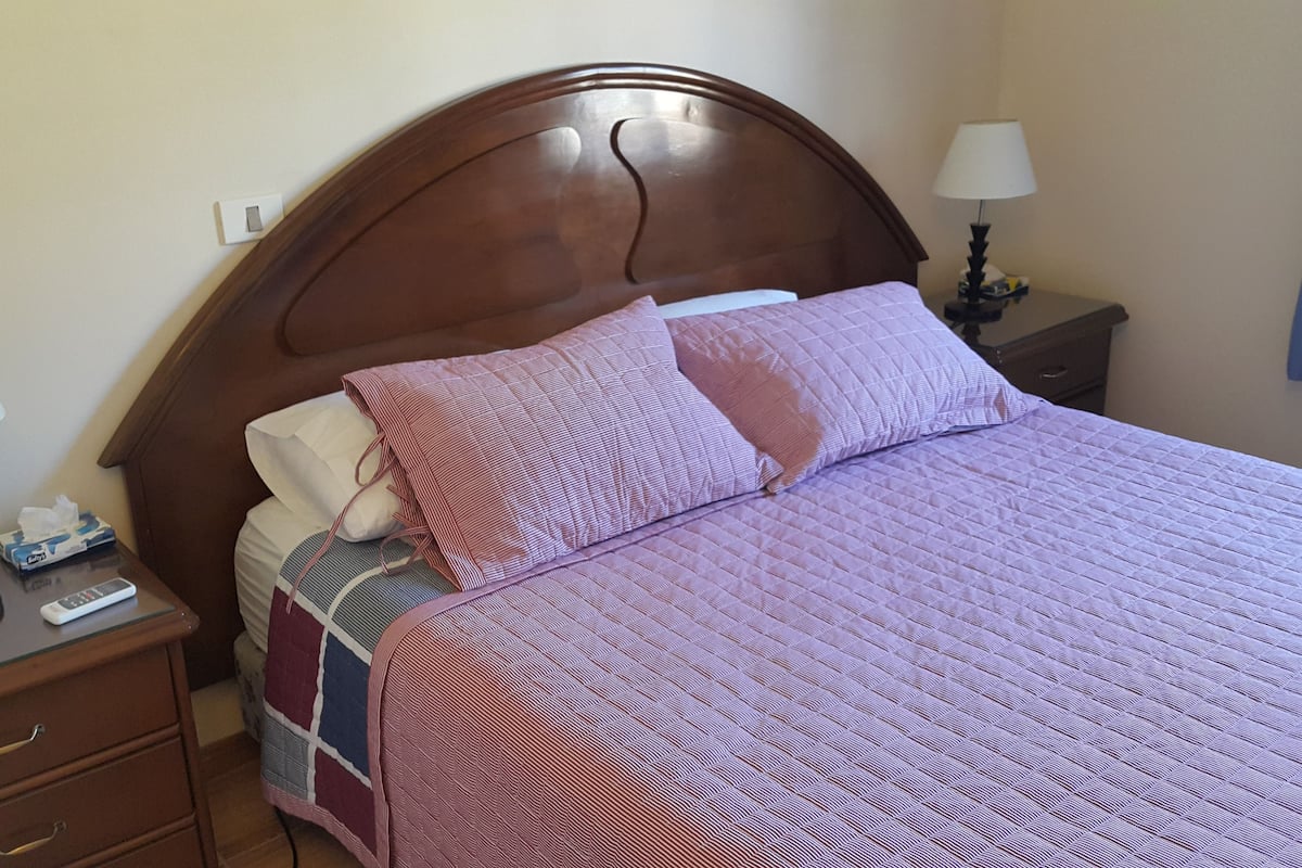 Habitación de hotel con cabecera de madera, cama con cubrecama acolchado, mesita de noche con lámpara y otro mueble auxiliar con lámpara.