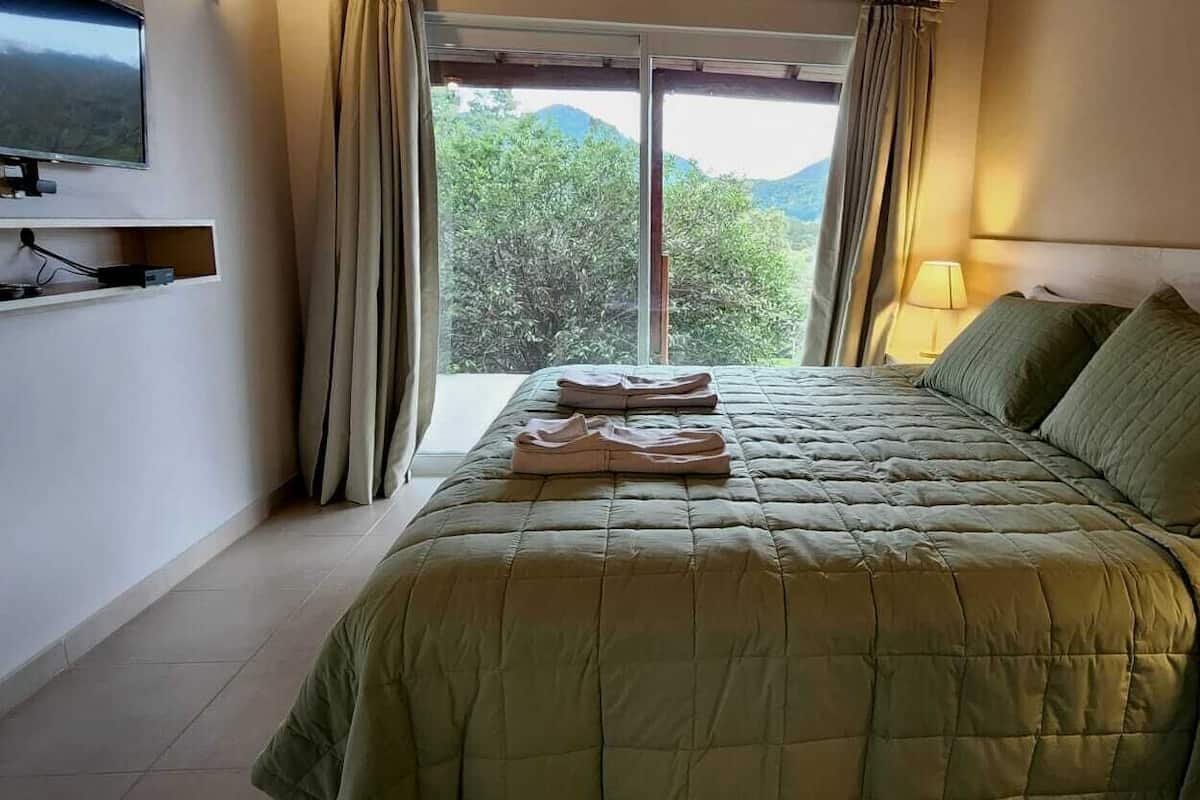 Habitación de hotel con cama, televisor montado en la pared, ventana con cortinas y lámpara en la mesita de noche.