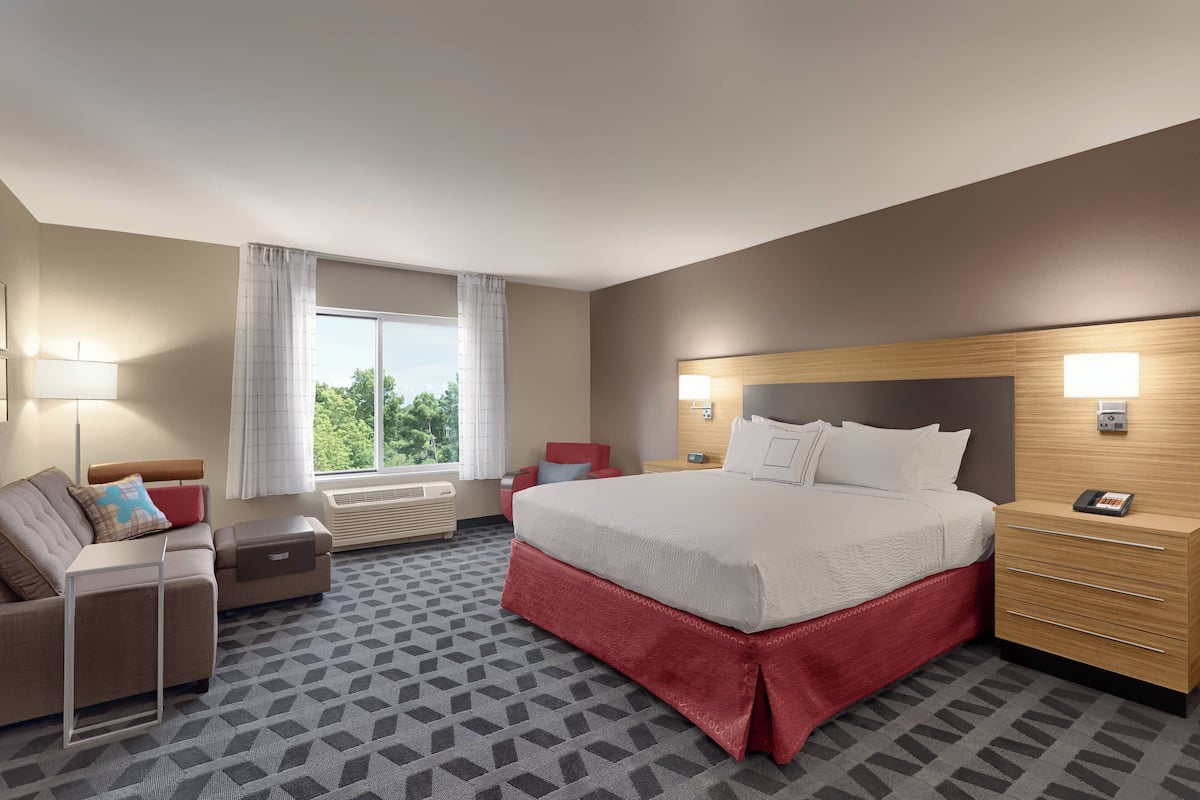 A hotel room with a large bed, a desk, a chair, a sofa, a window with curtains, and a patterned carpet.