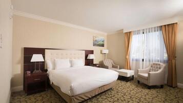 A hotel room with a large bed, a bedside table with a lamp, a chair, a small table, and a window with curtains.