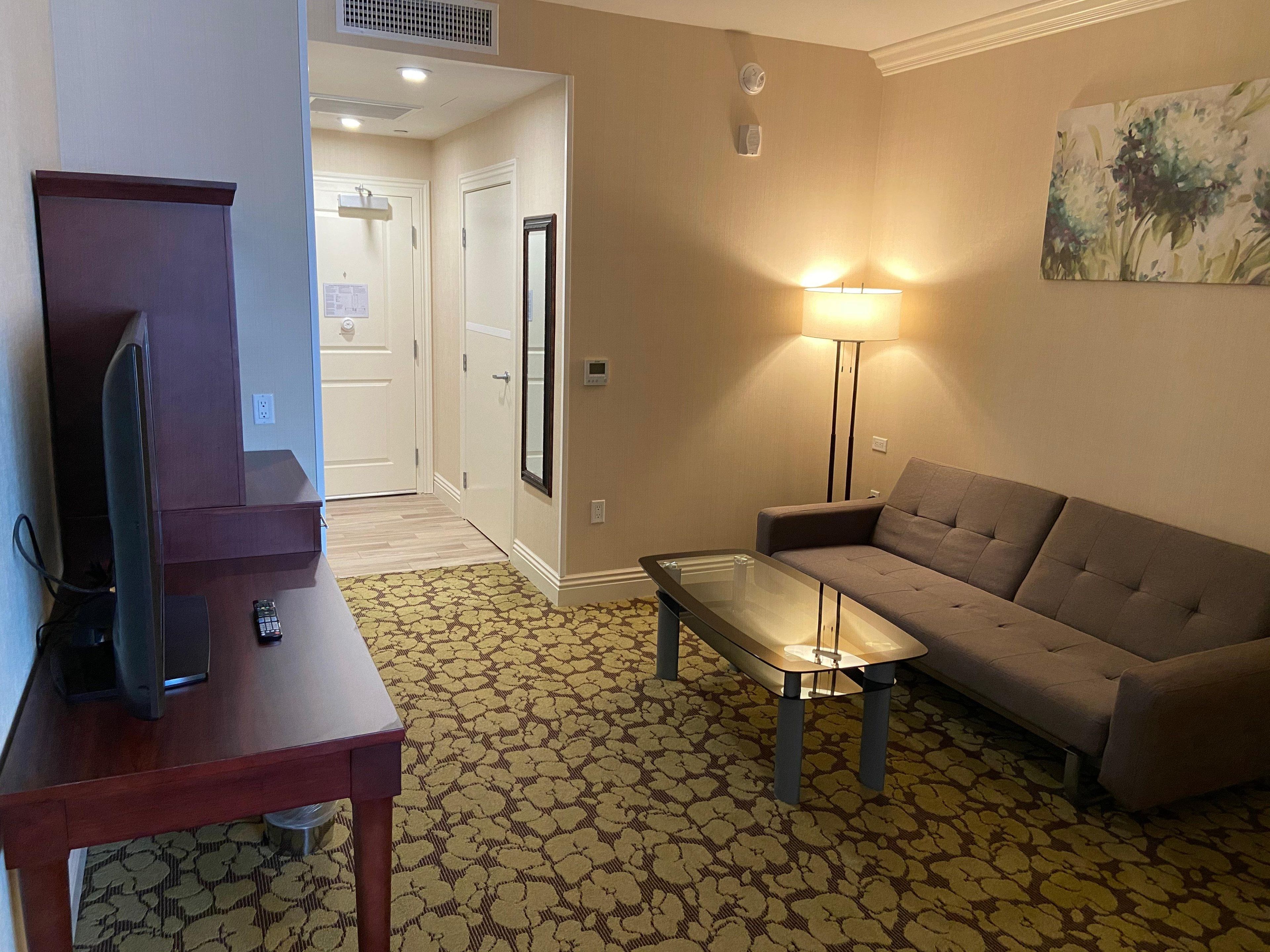 A hotel room with a sofa, a small table, a TV on a stand, a lamp, and a patterned carpet.
