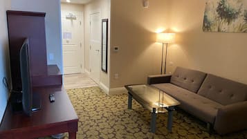 A hotel room with a sofa, a small table, a TV on a stand, a lamp, and a patterned carpet.