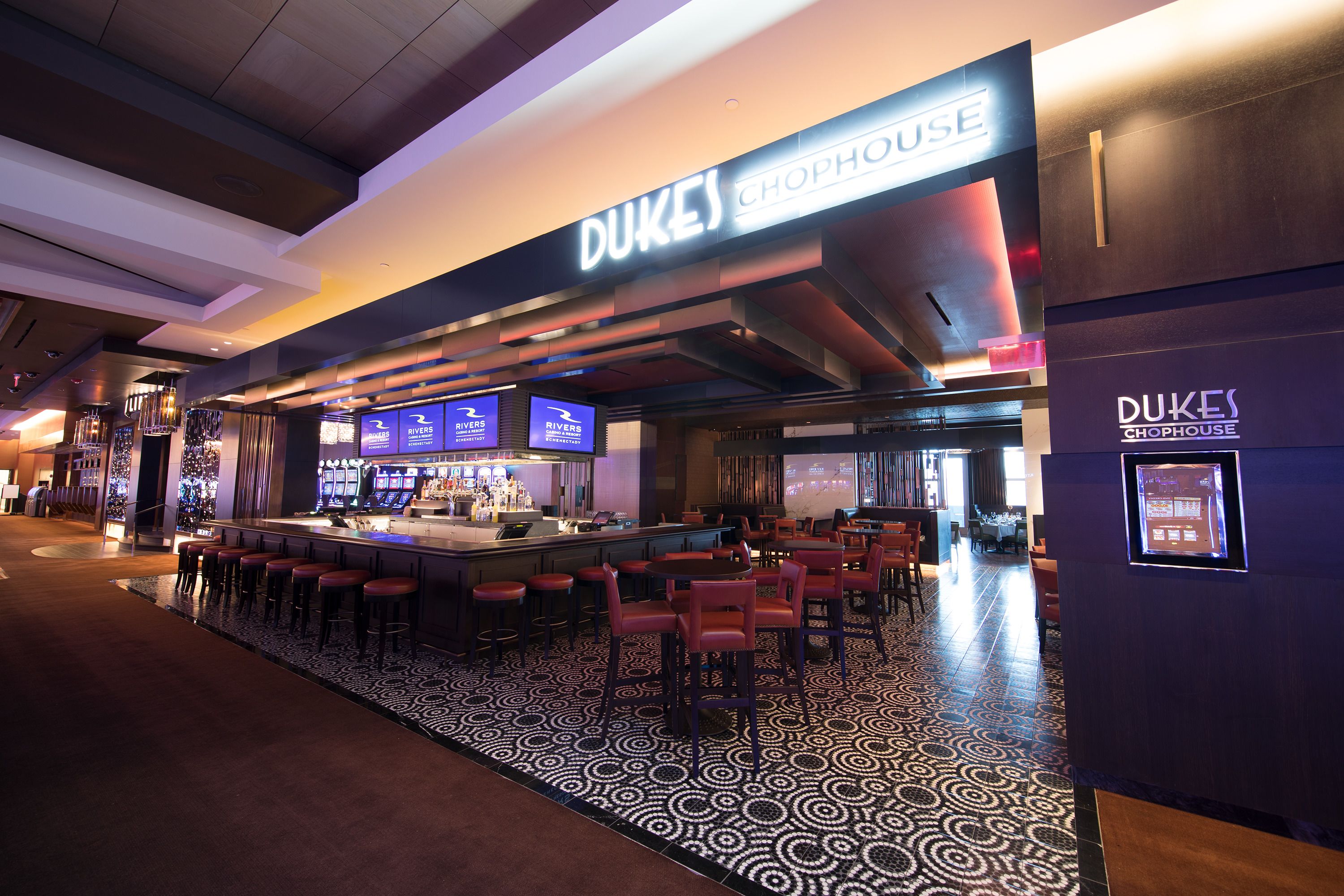 A bar area with a counter and multiple stools, a patterned floor, and a sign reading "DUKES CHOPHOUSE".
