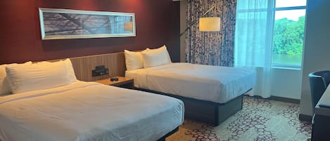 A hotel room with two beds, a painting on the wall, a lamp, and a window with curtains.