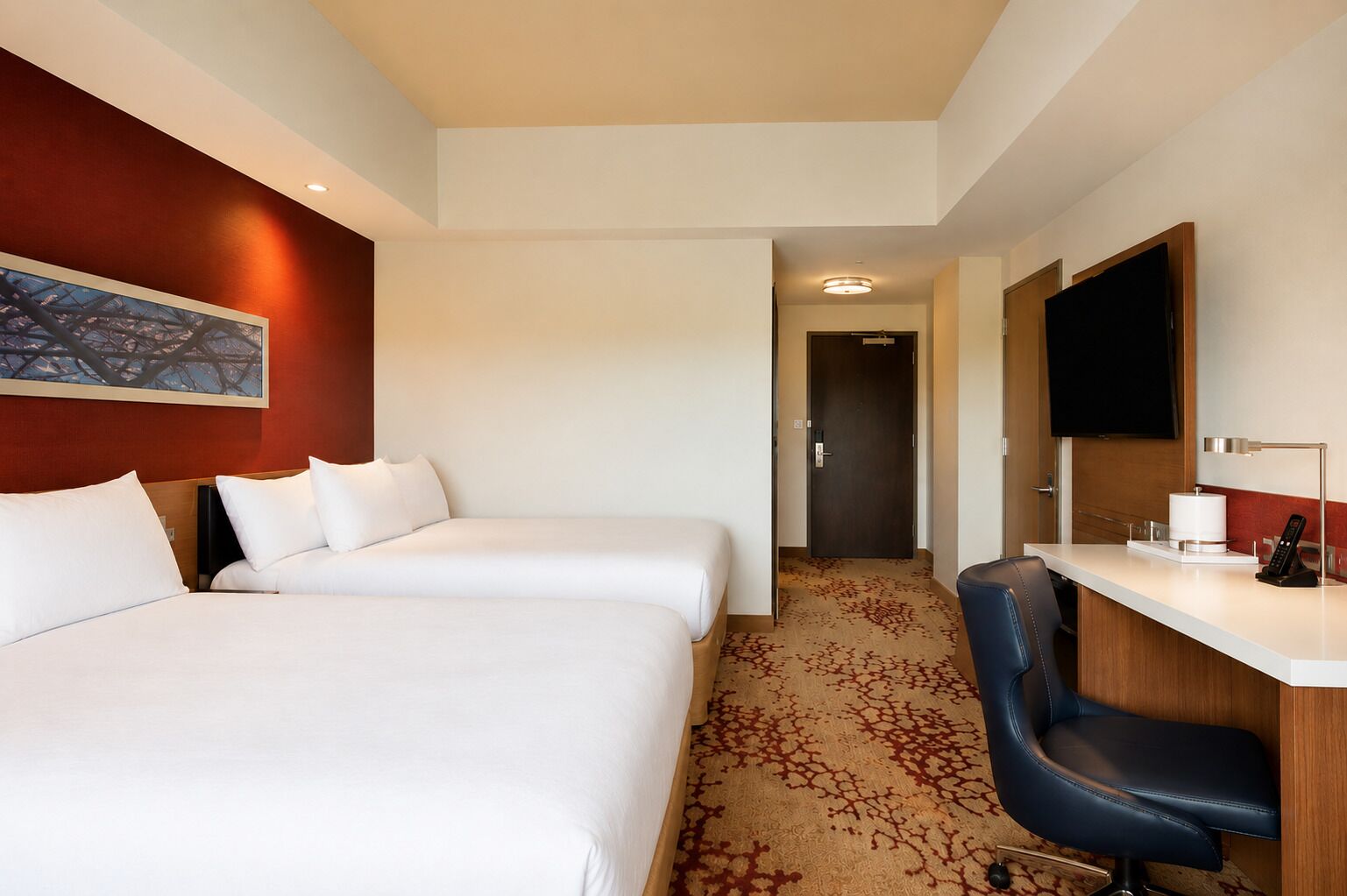 A hotel room with two beds, a desk with a chair, a flat-screen TV, and a painting on the wall.