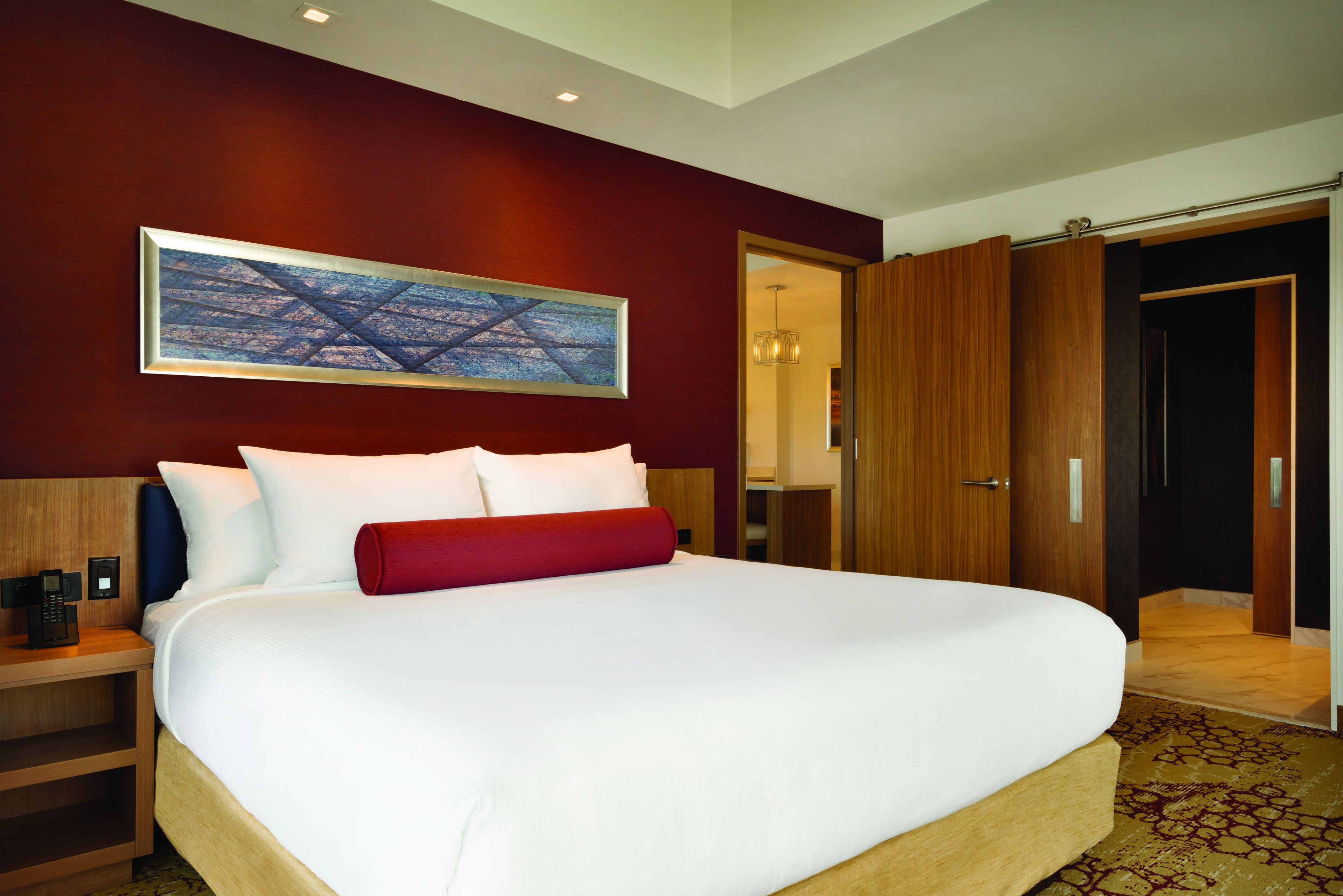 A large bed with white and red bedding, a wooden headboard, a nightstand with a phone, and a painting on the wall.