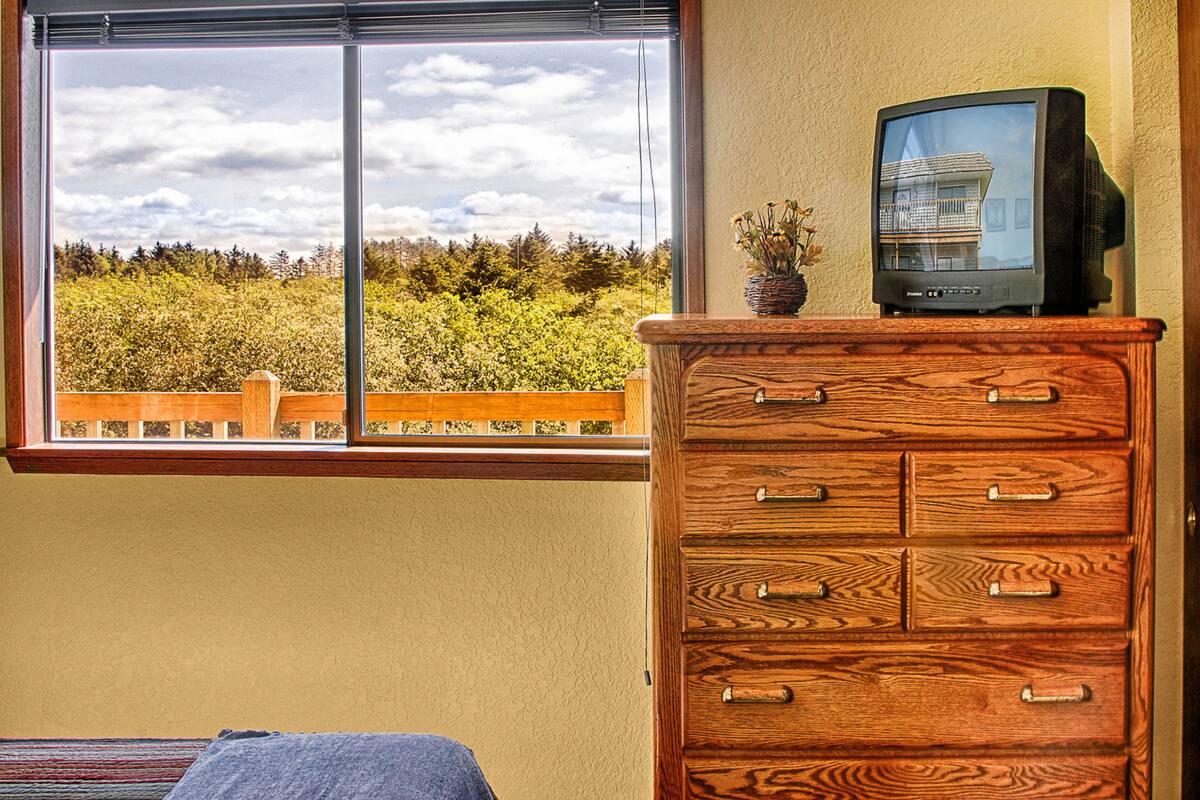 A room with a wooden dresser, a television on top, and a window with a view of trees and a cloudy sky.