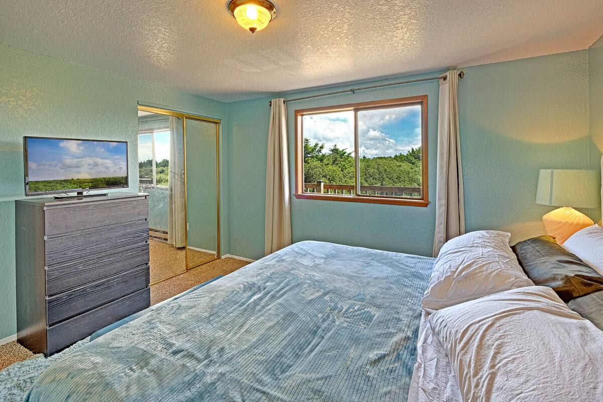 A bedroom with a bed, a dresser, a TV, a window with a view of greenery, and a ceiling light.