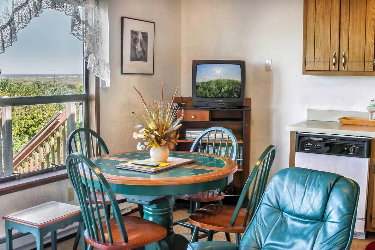 A dining area with a round table, chairs, a vase with flowers, a television, and a dishwasher.