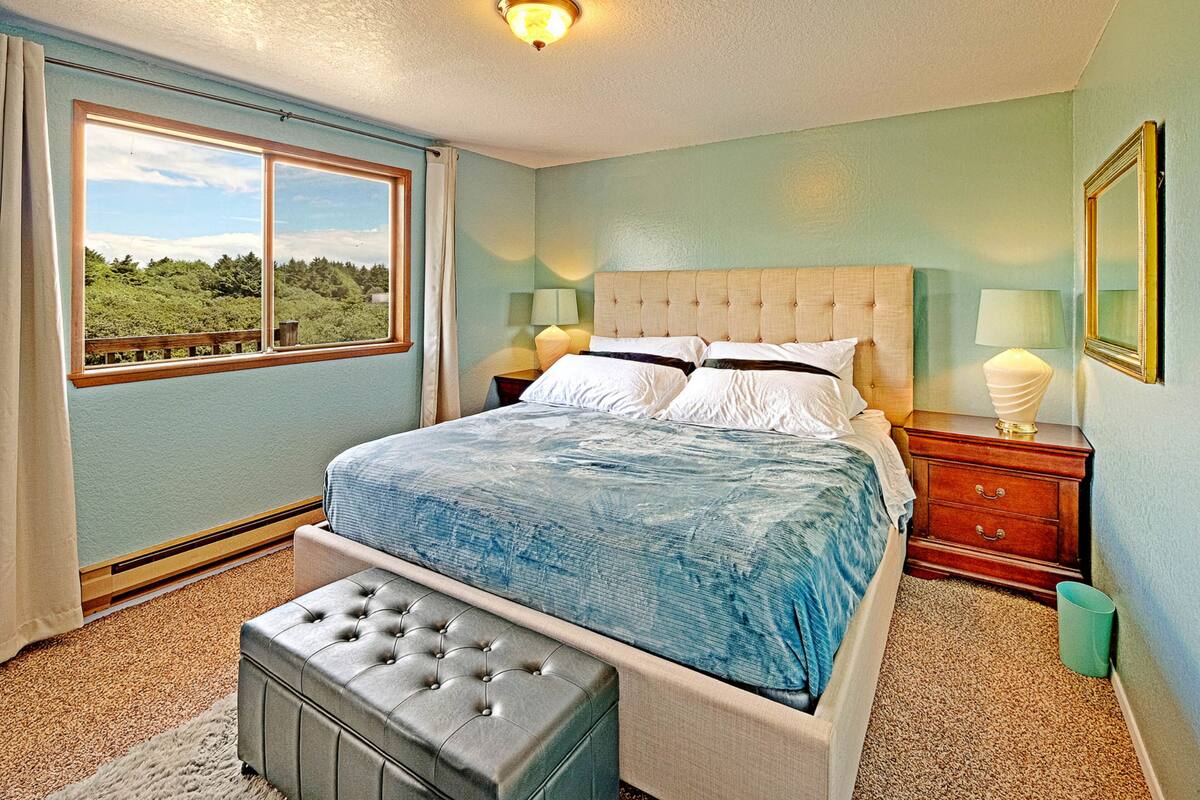 A bedroom with a large bed, two bedside tables, a window with a view, and a green trash bin.