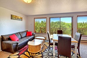 A living room with a black leather sofa, red cushions, a wooden coffee table, and a dining table with chairs. Large windows offer a view of trees and a wooden deck. - Ocean Surf Condos (Ocean Shores)