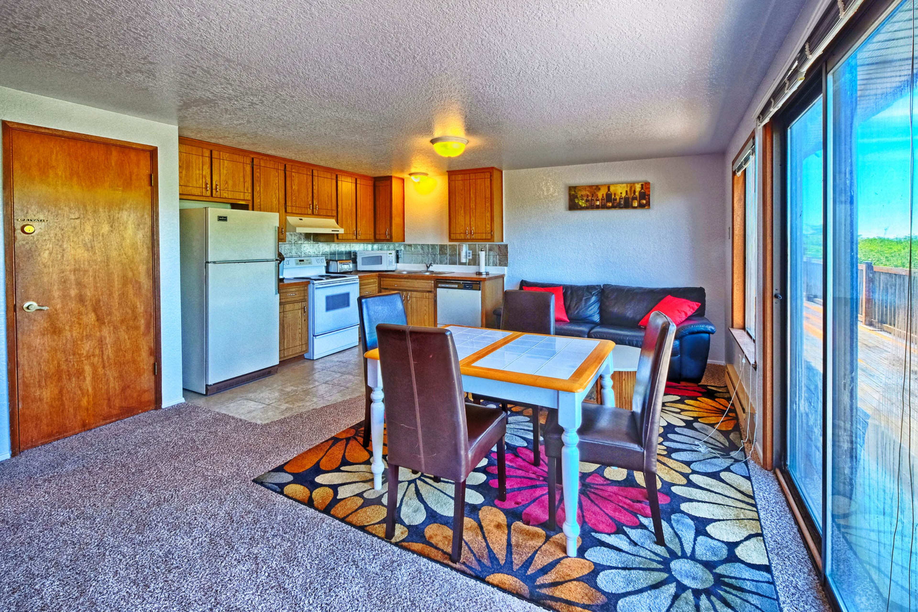 A compact kitchen with wooden cabinets, a refrigerator, and a microwave. There is a dining area with a table and chairs, and a sofa with a red cushion. The room has a large window providing a view of the outdoors.
