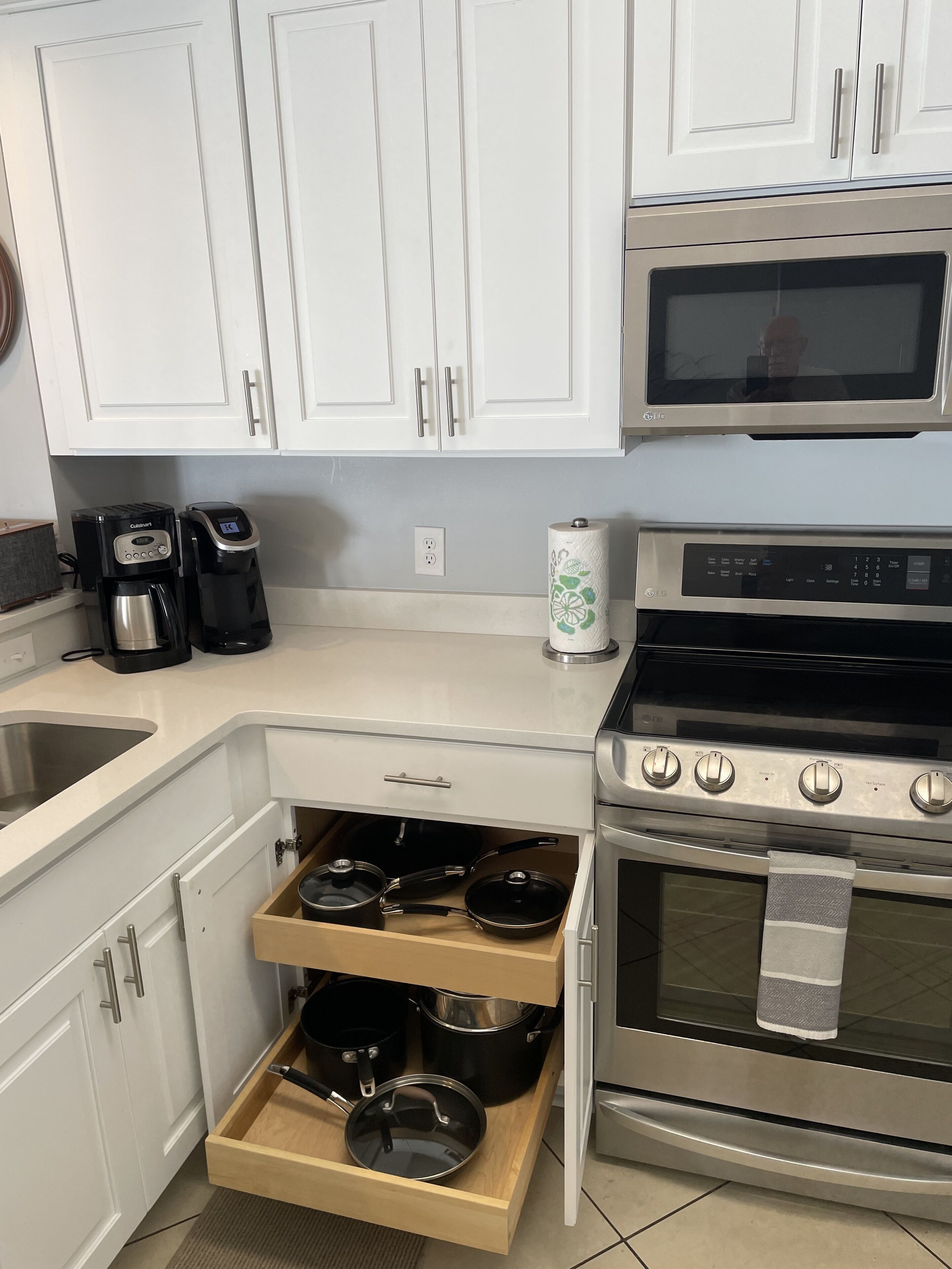 Una cocina con armarios blancos, un microondas de acero inoxidable, una cafetera, una estufa y un cajón con ollas.