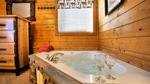 A wooden cabin interior with a bathtub, two wine glasses, a wooden dresser, and a window with blinds.