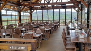 A spacious outdoor dining area with wooden tables and chairs, large windows offering mountain views, and a sign indicating restrooms.