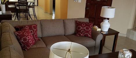 Ein Wohnzimmer mit einer Couch, einem Tisch mit Lampe und einer Küche im Hintergrund.