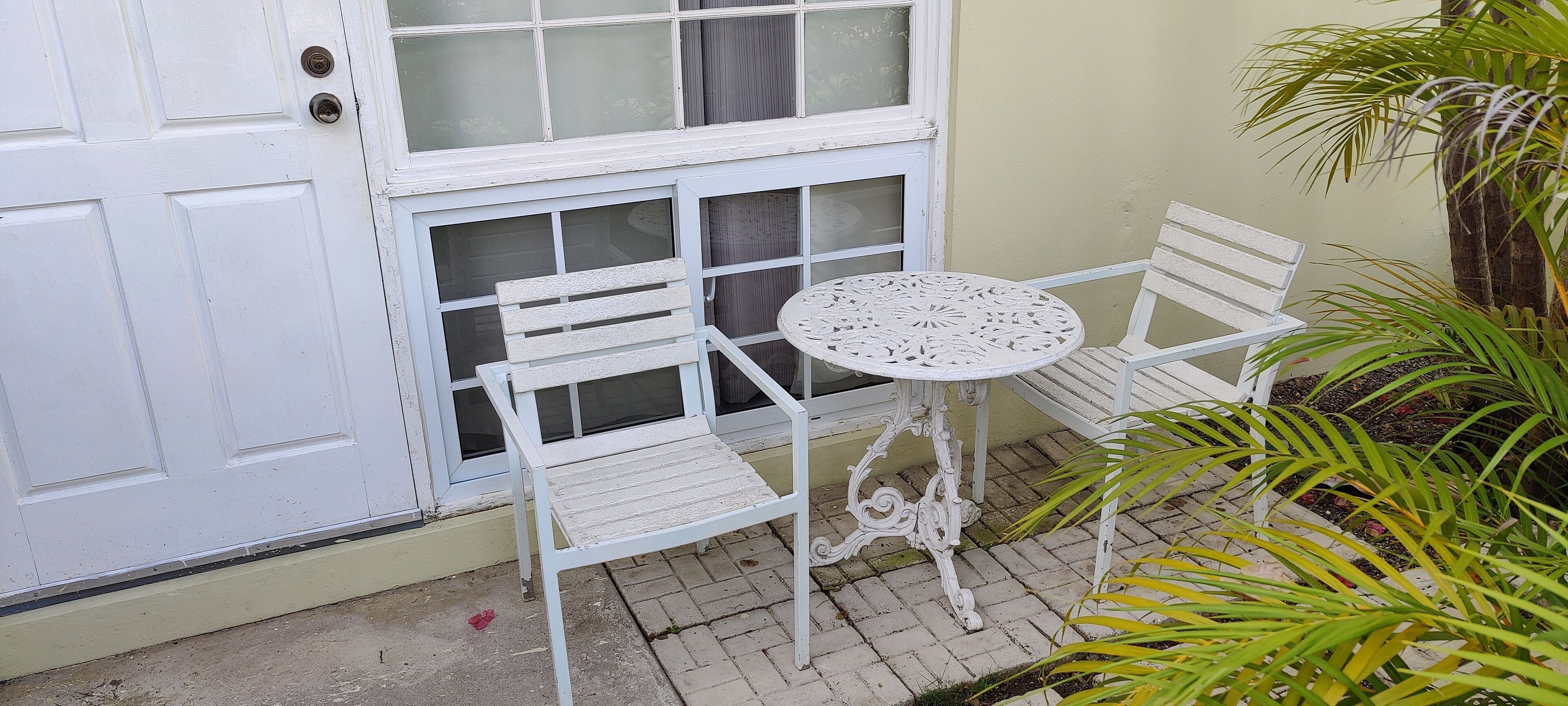 Een witte deur met een raam, een witte tafel en twee witte stoelen in een klein buitengebied met een betegelde vloer en een plant.