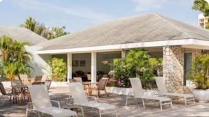 A covered outdoor seating area with lounge chairs and tables, surrounded by greenery and a stone wall.