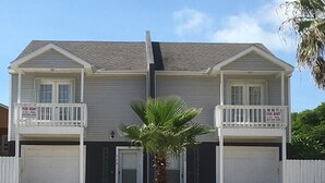Une maison à deux étages avec un garage à l’avant, un petit balcon au premier étage et un palmier dans le jardin.