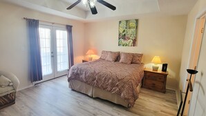 Une chambre à coucher avec un lit, deux tables de chevet avec des lampes, un ventilateur de plafond, une porte avec des rideaux et un mur orné d’un tableau.