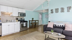 Una cocina con armarios blancos, una estufa y un microondas. Hay una zona de comedor con una mesa y sillas. Un sofá se encuentra en la sala de estar con una planta encima. En la pared se lee la palabra 'SURF'.