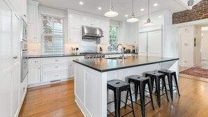 Una cocina con armarios blancos, una isla central con taburetes, un extractor de acero inoxidable e iluminación colgante.